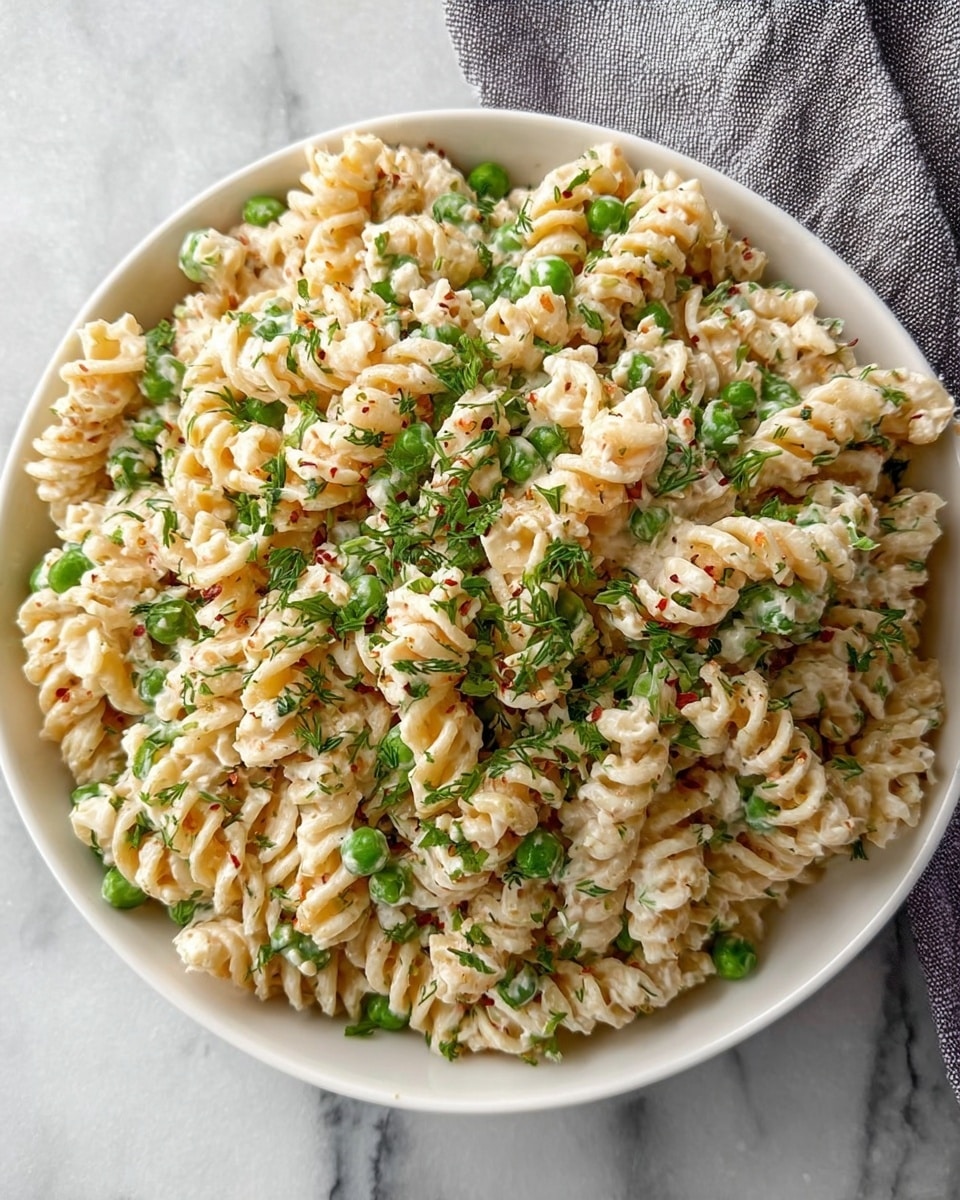 A white bowl filled with spiral rotini pasta mixed evenly with small green peas and creamy white sauce with visible herbs and small bits of seasoning. The pasta is coated well in the thick sauce, giving a smooth and slightly glossy texture. The dish is sprinkled with finely chopped green herbs and a light dusting of red pepper flakes and dried dill on top, adding color contrast. The bowl sits on a white marbled surface with a grey cloth napkin partly visible at the top right corner. The overall look is fresh and creamy with green and red specks over the light beige pasta photo taken with an iphone --ar 4:5 --v 7