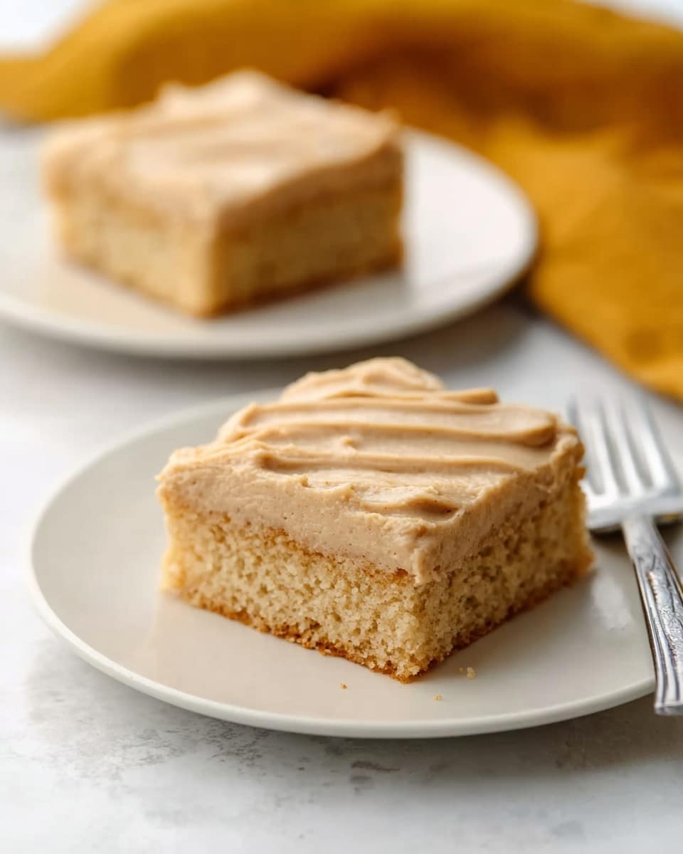 The image shows a square piece of cake with smooth, light brown frosting on top, arranged with soft ridges. The cake layer beneath the frosting is tan and looks moist, with a slightly crumbly texture. The cake is placed on a simple white plate set on a white marbled surface. In the background, there is a second square piece of the same cake on another white plate, slightly out of focus. A metal fork lies to the right side of the closest plate, and a mustard-yellow cloth is softly blurred behind, adding a warm tone to the scene. Photo taken with an iphone --ar 4:5 --v 7