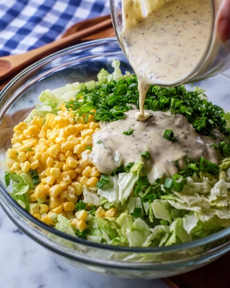 A clear glass bowl on a white marbled surface holds a colorful layered salad. At the bottom, finely chopped light green lettuce fills most of the bowl. To one side, bright yellow corn kernels form a small mound. Dark green chopped leafy herbs sit next to the lettuce, with finely sliced green onions beside them. A woman's hand is pouring a creamy, pale dressing thick with specks over the top, draping over the greens and corn in smooth streams. The scene includes a wooden spoon and a blue and white check cloth in soft focus in the background. Photo taken with an iphone --ar 4:5 --v 7