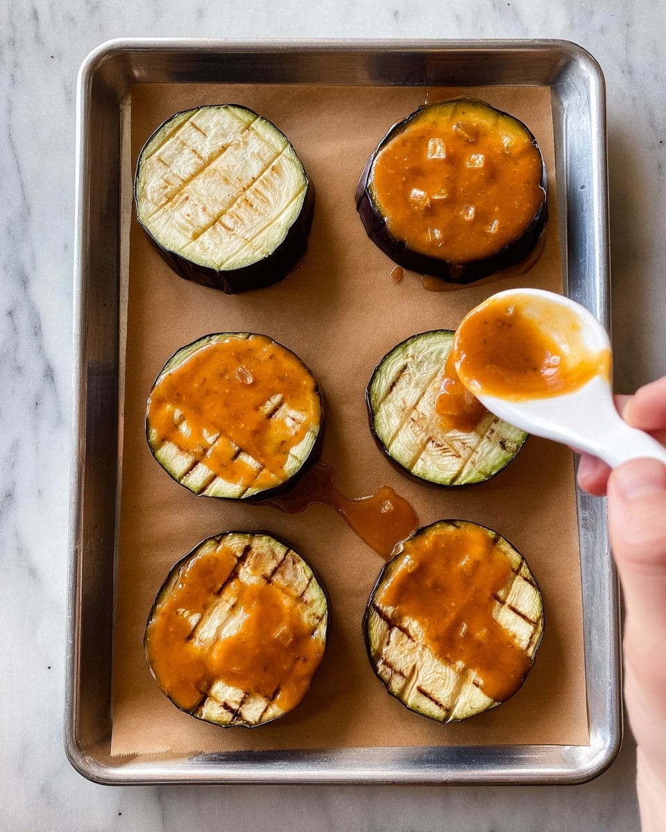 A silver baking tray lined with brown parchment paper holds six thick, round slices of eggplant arranged in two rows. Each eggplant slice has a dark purple outer skin and a pale, creamy interior with visible crisscross cuts on top. Four of the slices have a shiny, thick orange sauce spread unevenly on top, with small chunks visible in the sauce. A woman's hand is seen holding a white ceramic spoon, pouring the same orange sauce onto one of the plain eggplant slices. All of this is set against a white marbled surface. photo taken with an iphone --ar 4:5 --v 7