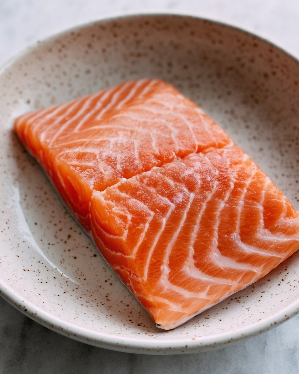 A piece of fresh raw salmon fillet with a smooth, shiny orange surface and light white stripes lies flat in the middle of a white bowl with small speckles, the bowl placed on a white marbled surface. The salmon shows soft, moist texture with fine lines running parallel on both sides. Photo taken with an iphone --ar 4:5 --v 7
