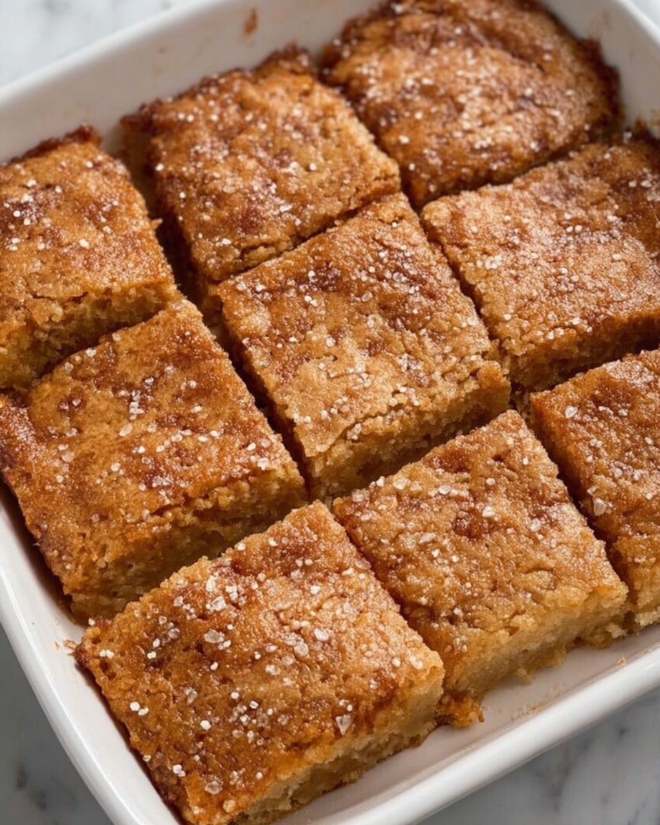 This image shows a white rectangular dish filled with nine square pieces of a brown baked dessert. Each piece has a slightly rough texture with a light dusting of sugar crystals on top. The dessert appears soft and slightly fluffy with a golden-brown color. The squares are neatly separated with visible lines between them, and the dessert fills the entire dish. The dish is set on a white marbled surface. photo taken with an iphone --ar 4:5 --v 7